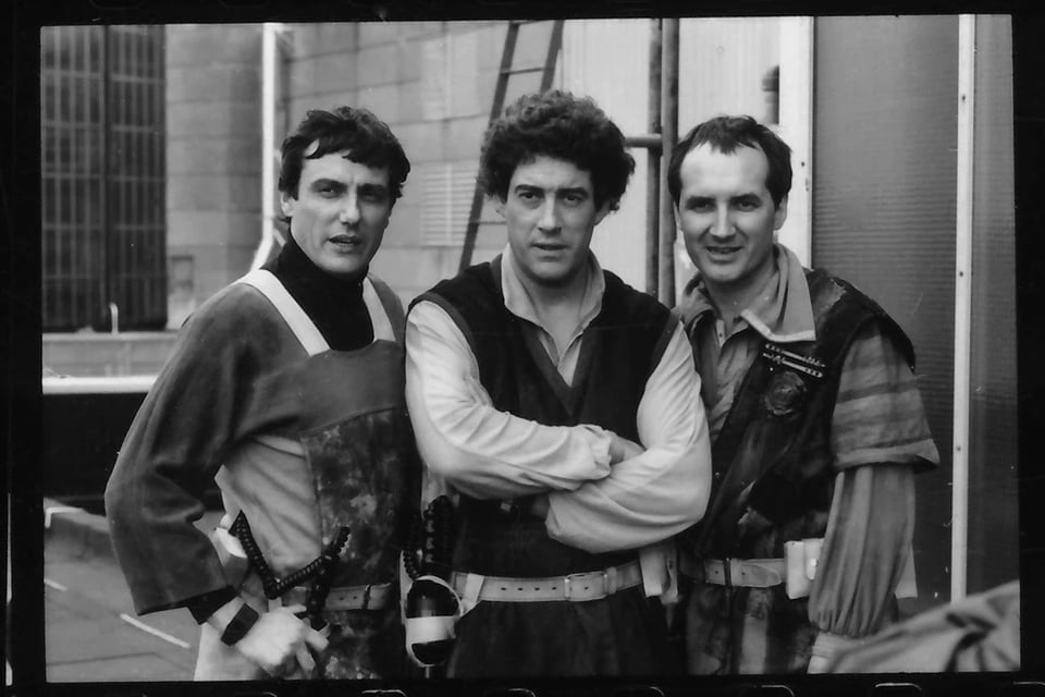 Three men stand in front of a cement wall with a ladder. They all have puffy sleeves and big collars. The man on the left is wearing a kind of smock and holding onto a cord like an old-fashioned telephone cord. The man in the middle has curly hair and folded arms.