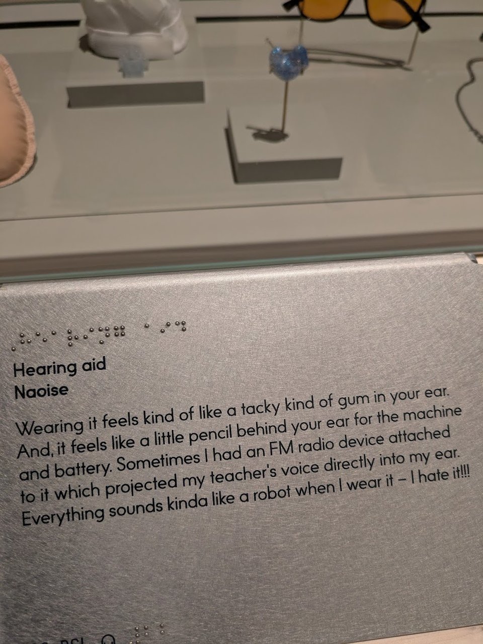 A case with multiple objects, including a blue sparkly earmold. The label says "Hearing aid" and quotes from the donor, apparently a child, about their experience wearing a hearing aid.