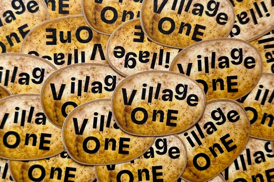 Photo of a pile of Village One logo stickers, all showing a potato texture with the words “Village One” on top