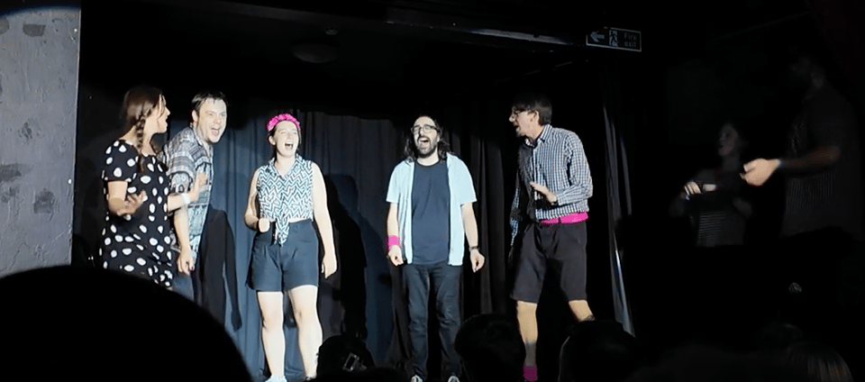 A group of five performers stands on stage, smiling and engaging with the audience during a lively theatrical performance.