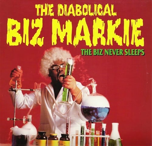 On a red background, a Black man is dressed like a stereotypical mad scientist: white lab coat, glasses, a cheap curly wig of white hair. Before him on a table are stereotypical mad scientist chemistry equipment: beakers and test tubes and erlenmeyer flasks full of colored liquid with white gas pouring out of them. The man is holding a green beaker full of green liquid and is exclaiming in surprise.