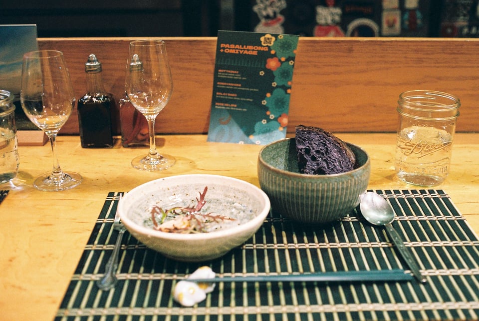 a scan of a color photo of a sashmi and ube bread dish sitting on a japanese placement on a wooden table with wine glasses at the back, and a menu for Ox and Tiger, a Filipino-Japanese restaurant in San Francisco
