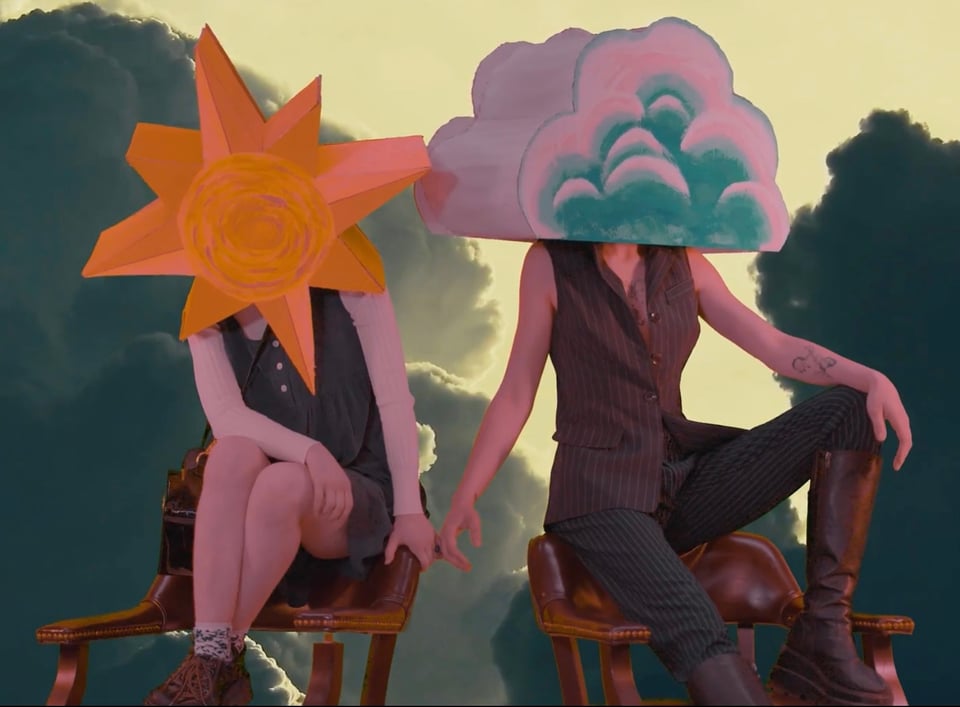 Two people are perched on top of the backs of two chairs, facing slightly away from one another. The person on the left (Copeland) is wearing a cardboard make shaped and painted like the sun, covering their face. The person on the right, Drew, is wearing a cardboard head shaped and painted like a cloud. Behind them is a greenscreen image of clouds.