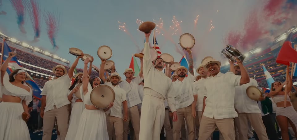 Bad bunny holds up a football to signal the end of his halftime show. He is surrounded by performers and dancers.