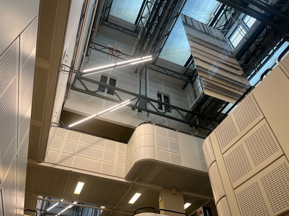 An interior view of a library space, looking up towards the ceiling. There are several balconies where people could stand, and black beams running through the ceiling space holding the roof up, far above where somebody could stand or reach.