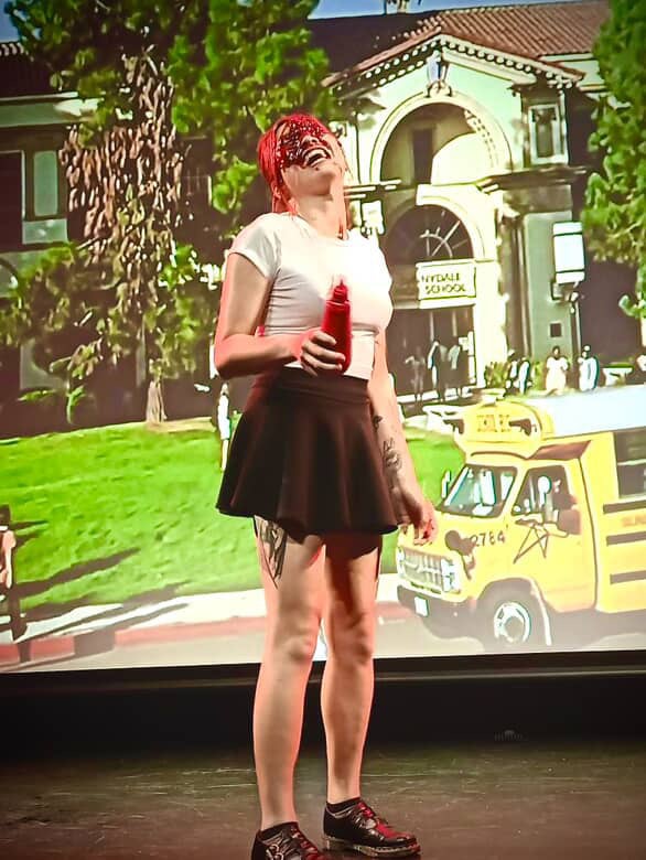 Lily Catastrophe standing in front of a high school backdrop in school uniform, holding a ketchup bottle, her eyes caked in ketchup blood