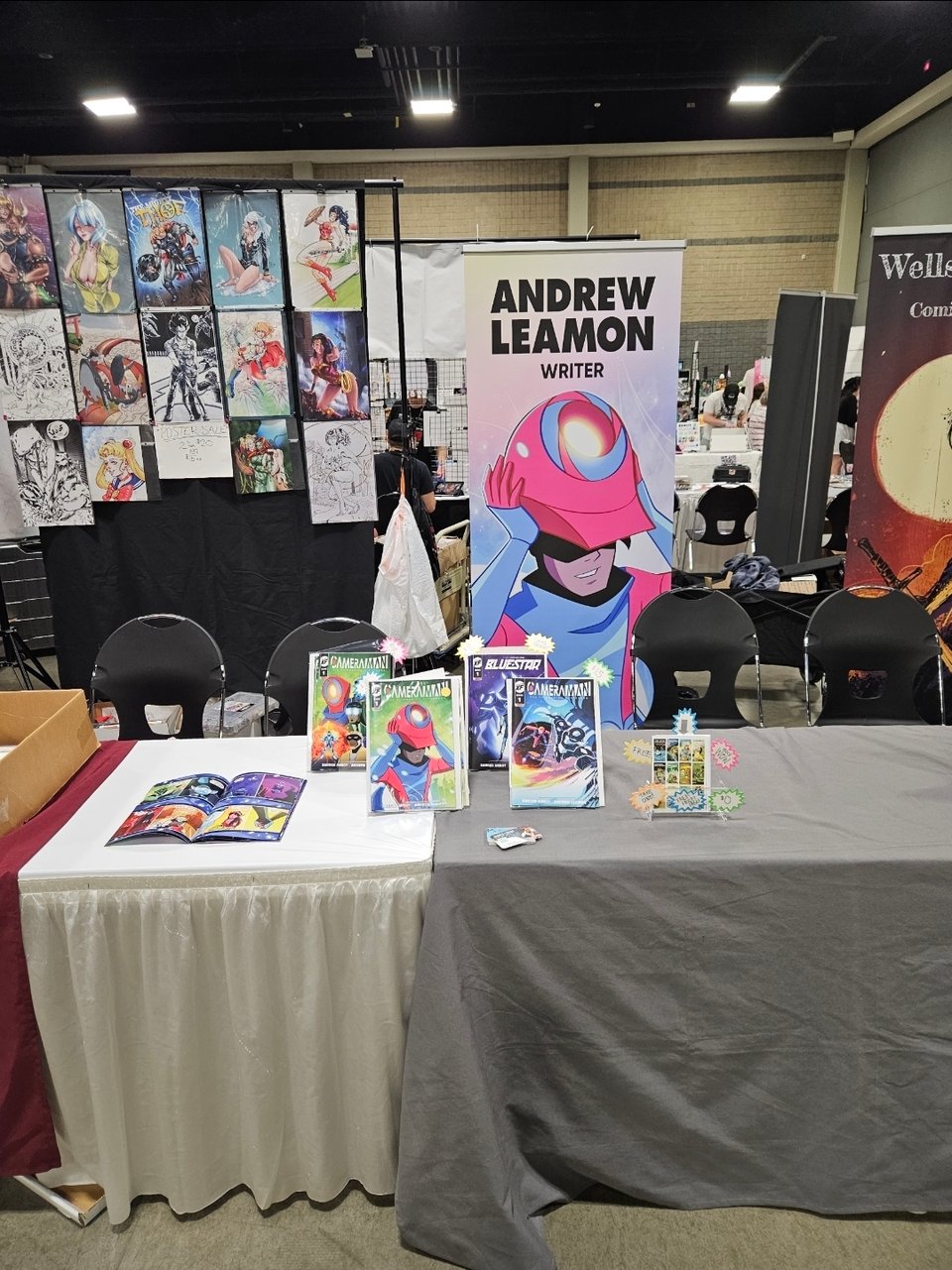 At a convention hall, a table with comics displayed on stands. Behind the table, a tall vertical sign that has the name ANDREW LEAMON and COMIC WRITER with an illustration of Camera Man.