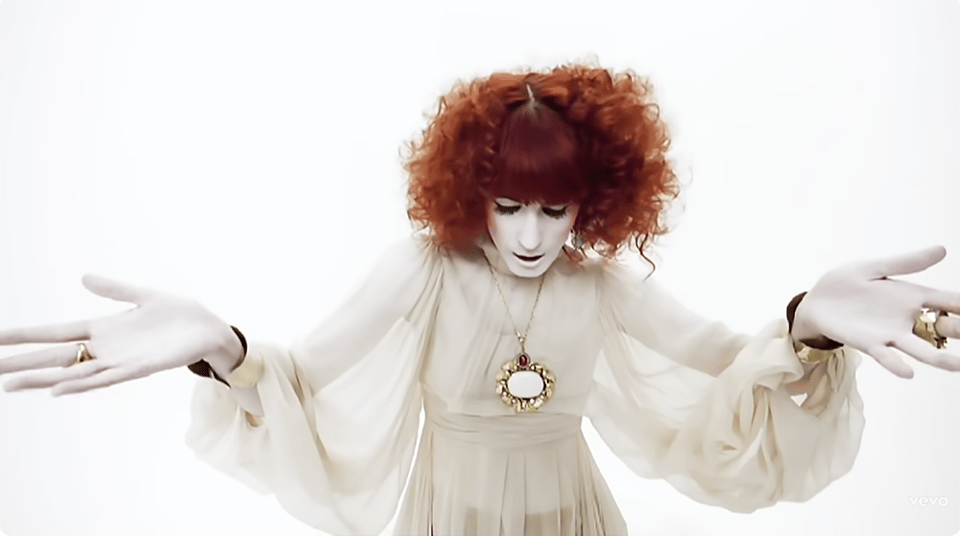Florence Welch of Florence and the Machine, from her video for Dog Days are Over, wearing a white gauzy gown, white makeup on her hands and face, a large mirrored gold pendant, and brilliant red curly hair.