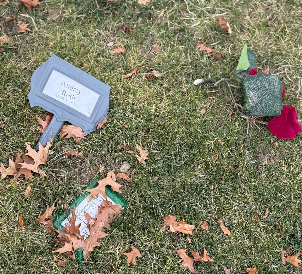 photo of grass with dried leaves and a temporary grave marker sticking out. The marker reads "Andrey Roth • 2/21/1938 - 7/10/2025"