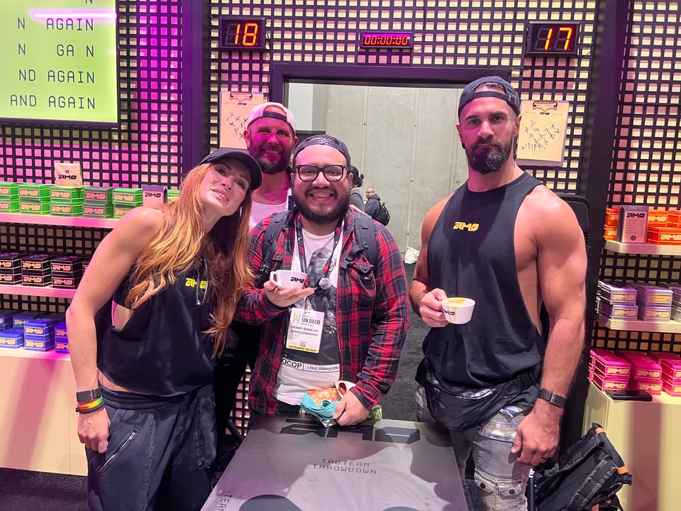 Three people at coffee convention. Henry standing between Becky lynch Seth Rollins and Josh Gallegos at the AMO booth