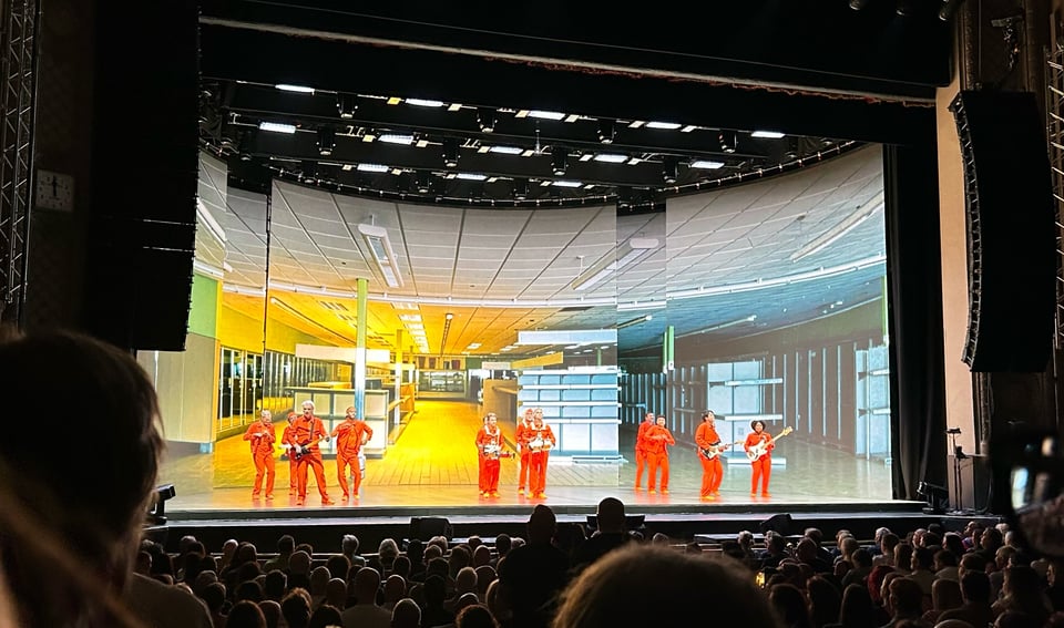 A group of musicians on a stage in front of an audience. They are all dressed in identical orange suits. There is a back projection of an empty office/server room behind them.