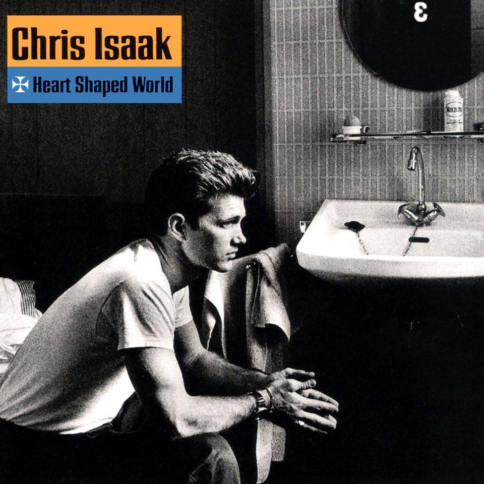 A black and white photo of Chris Isaak, a stupidly hansome white man, in a white t-shirt and dark pants. He's sitting on a bed in a motel room, his hands outstretched, fingertips touching. Behind him on the right side of the picture is a sink and mirror and some bath towels. He has a pensive expression on his face. It's a heart-shaped world, Chris.