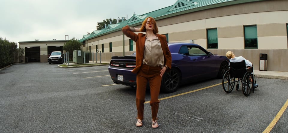 A still from Kinds of Kindness. Emma Stone dances in parking lot behind a purple car. A person in a wheelchair has their back to her.