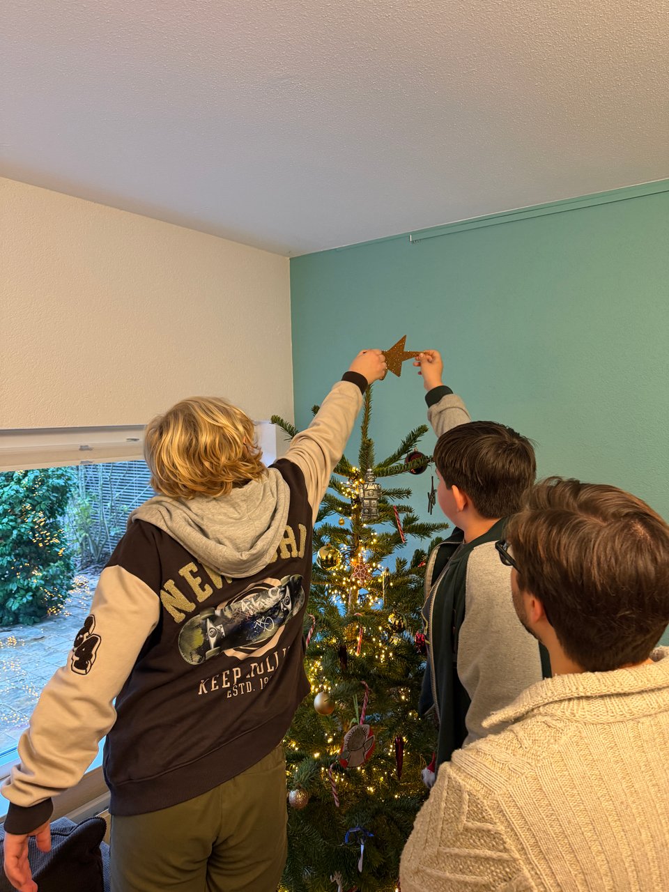 Two boys reach to the top of a Christmas tree to put on the star topper. A man looks on.