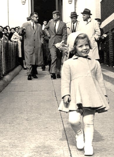 Young Caroline Kennedy gleefully goes for a stroll while her daddy behind her, the President of the United States, the most powerful person in all the world, carries her doll.