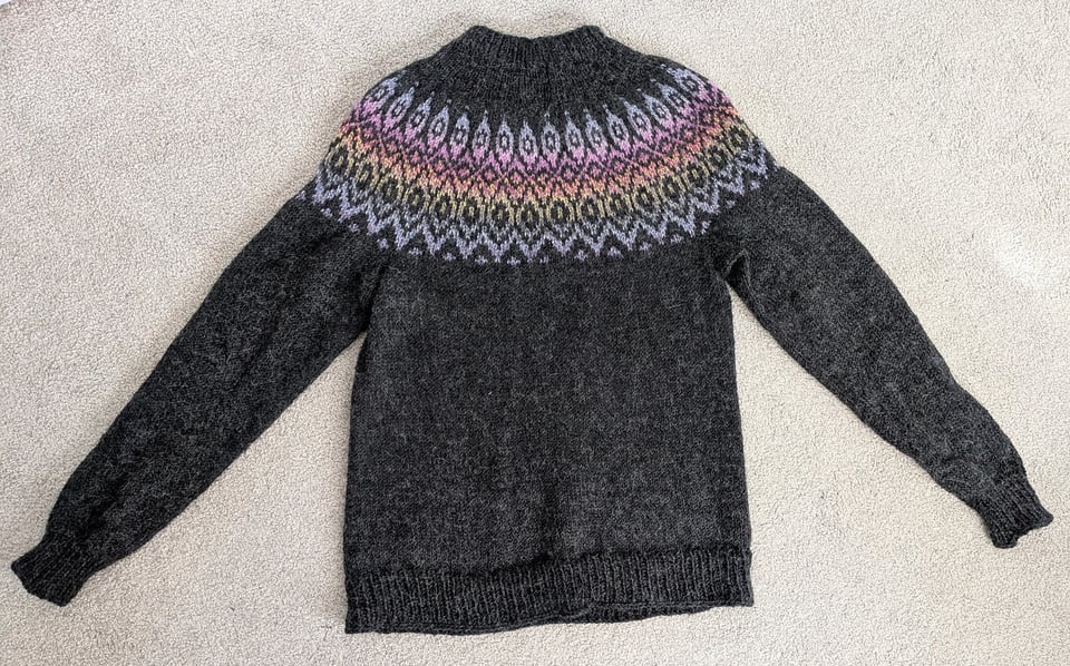 a dark charcoal grey sweater with a kind of diamond-patterned colorwork yoke in a gradient ranging from pale blue to pink to yellow to light grey and back to pale blue