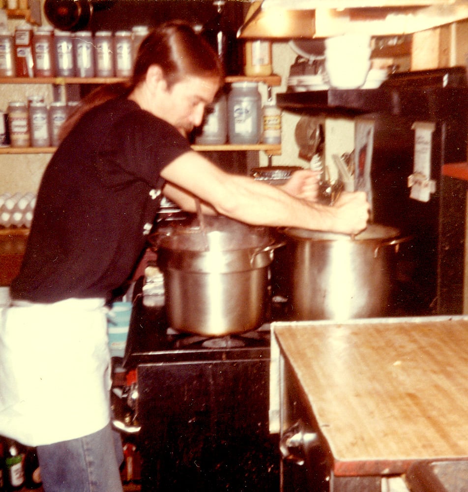 A man with a long ponytail, a black t-shirt and white apron stands in front of a restaurant-grade stove at the Iron Horse Music Hall.