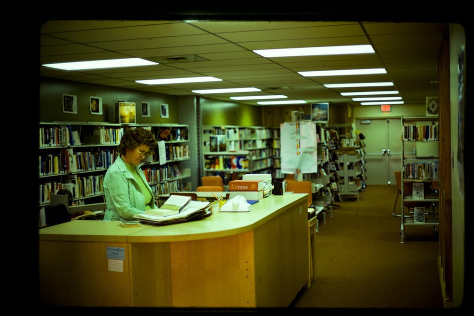 colour photo. A female librarian stands behind a desk. There are stacks of books in the background