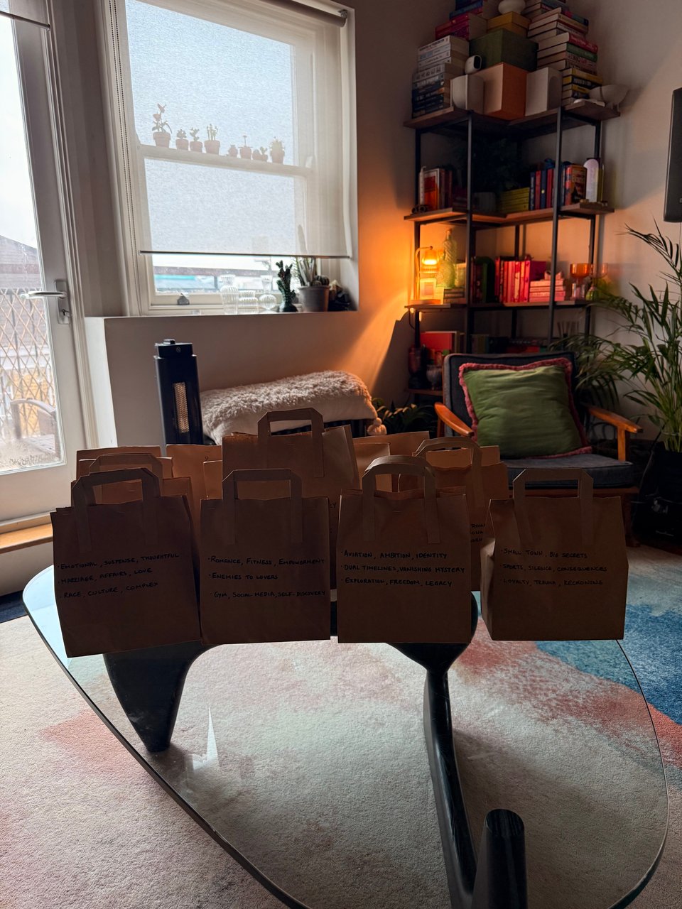 A cozy reading corner in an apartment featuring a mystery book swap with around 20 brown paper bags with handwritten notes arranged on a reflective coffee table. Behind the bags is a comfortable seating area with a green pillow, and a black bookshelf filled with colorful books illuminated by warm lighting. A window with small potted plants on the sill allows natural light into the space. The room has a colorful area rug with pink tones.