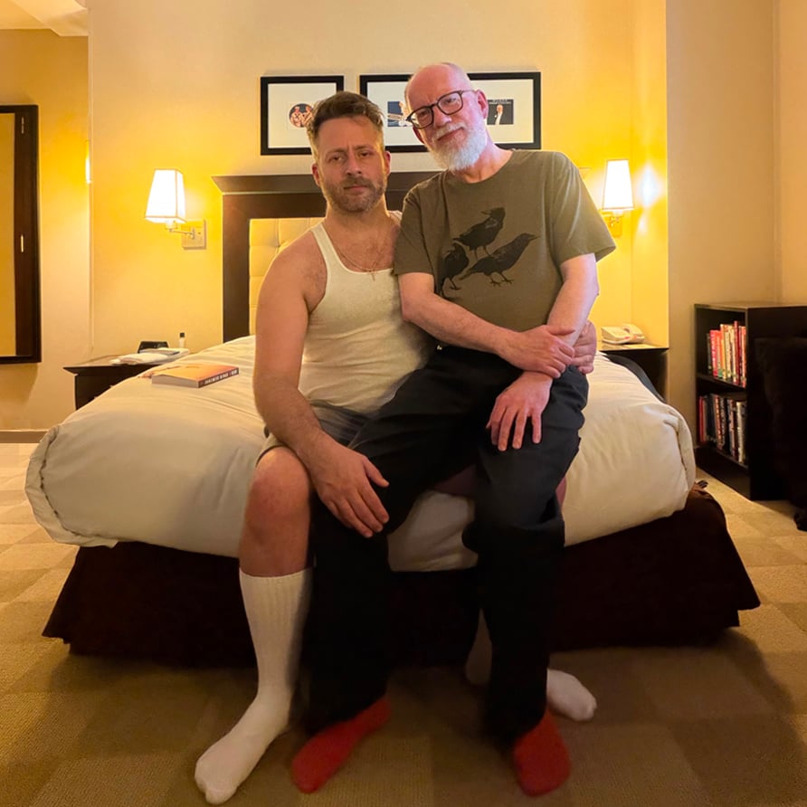 David sits on the lap of Joel Someone at the foot of the bed in a yellow-lit hotel room. Joel is wearing a white tank top, grey shorts and white socks. David is wearing a sage t-shirt with three crows printed on the front, black carpenter's pants and red socks.