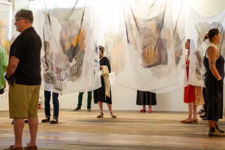 people walk through a white walled gallery, in which sheer printed textile art is hung from the ceiling like curtains