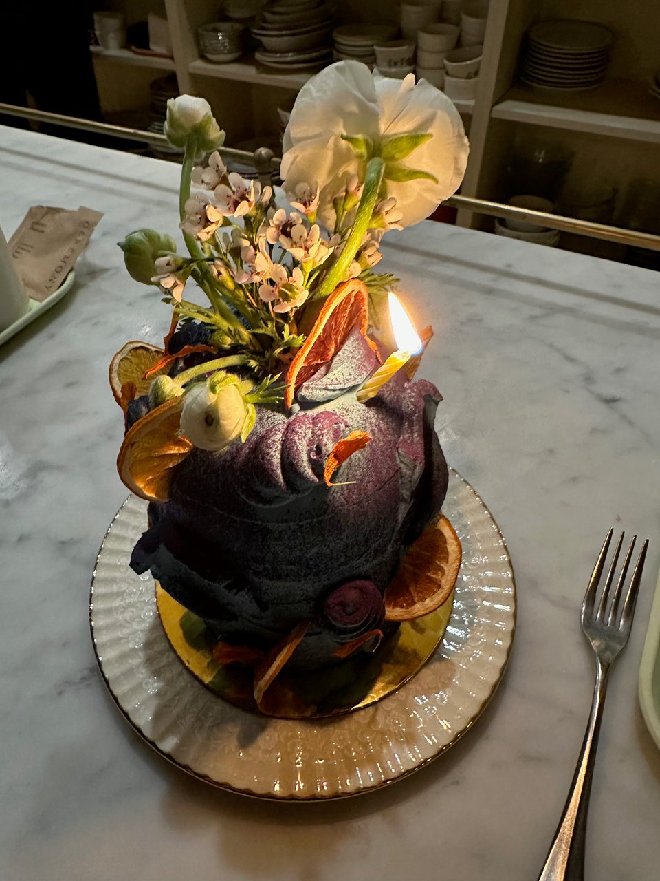 A moody, tall cake decorated with flowers, dried orange slices, and one lit candle sits on a small plate with fluted edges on a marble counter.