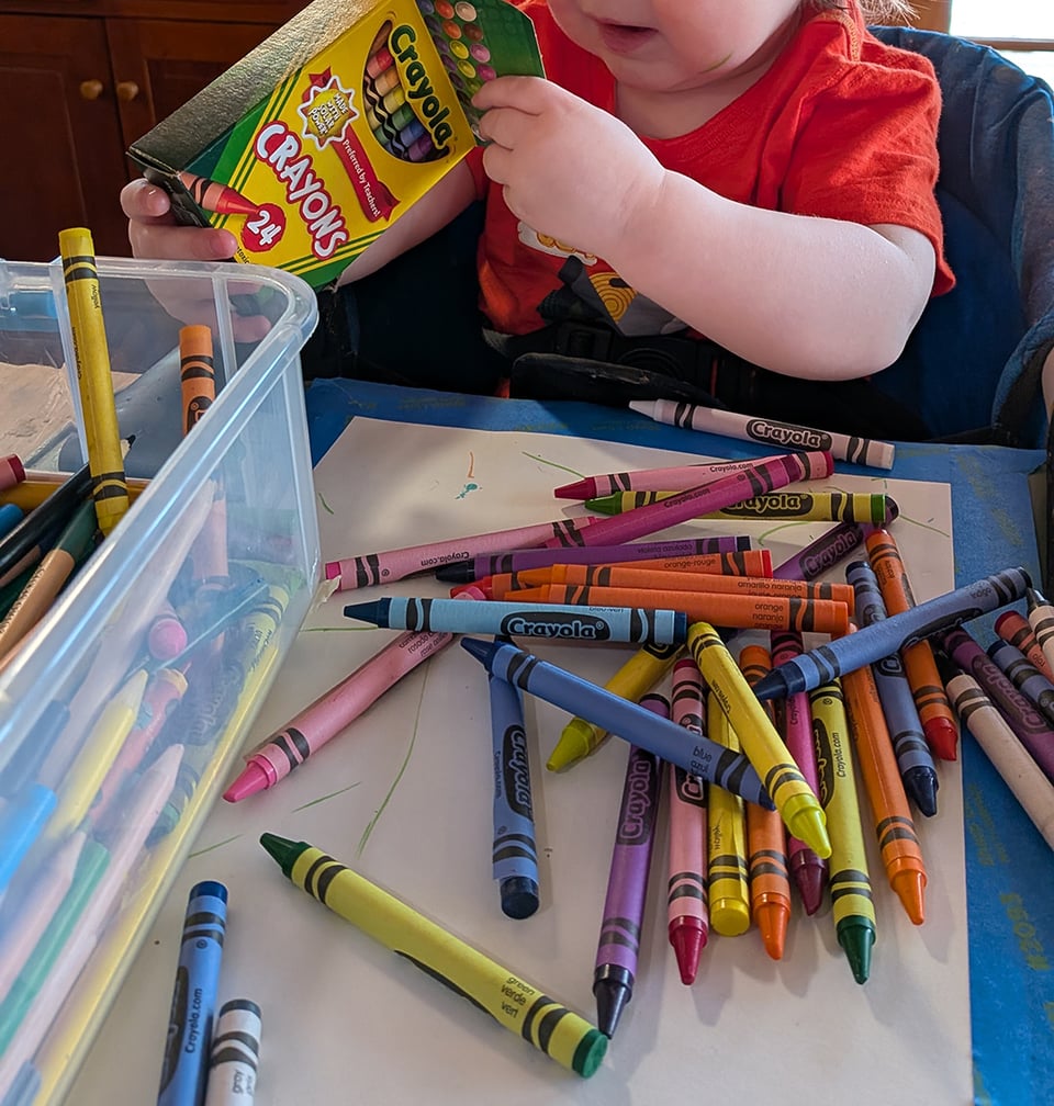 a full box of crayons is spilled over a piece of paper taped to the table; part of a young toddler's face can be seen as the child looks through the box to see if there are any more crayons. It's chaos.