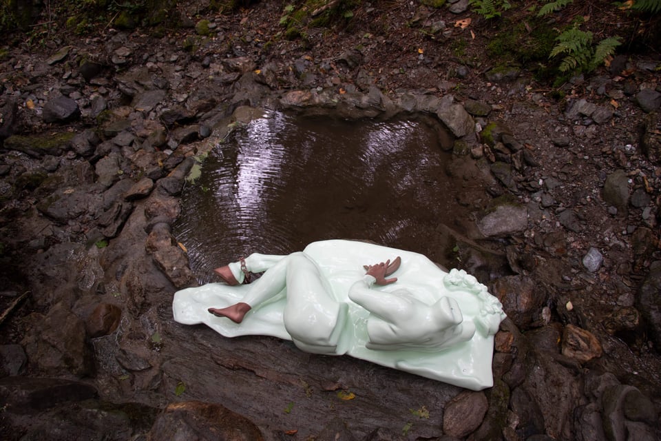 A figure that is also a fountain reclining by a pool in the forest