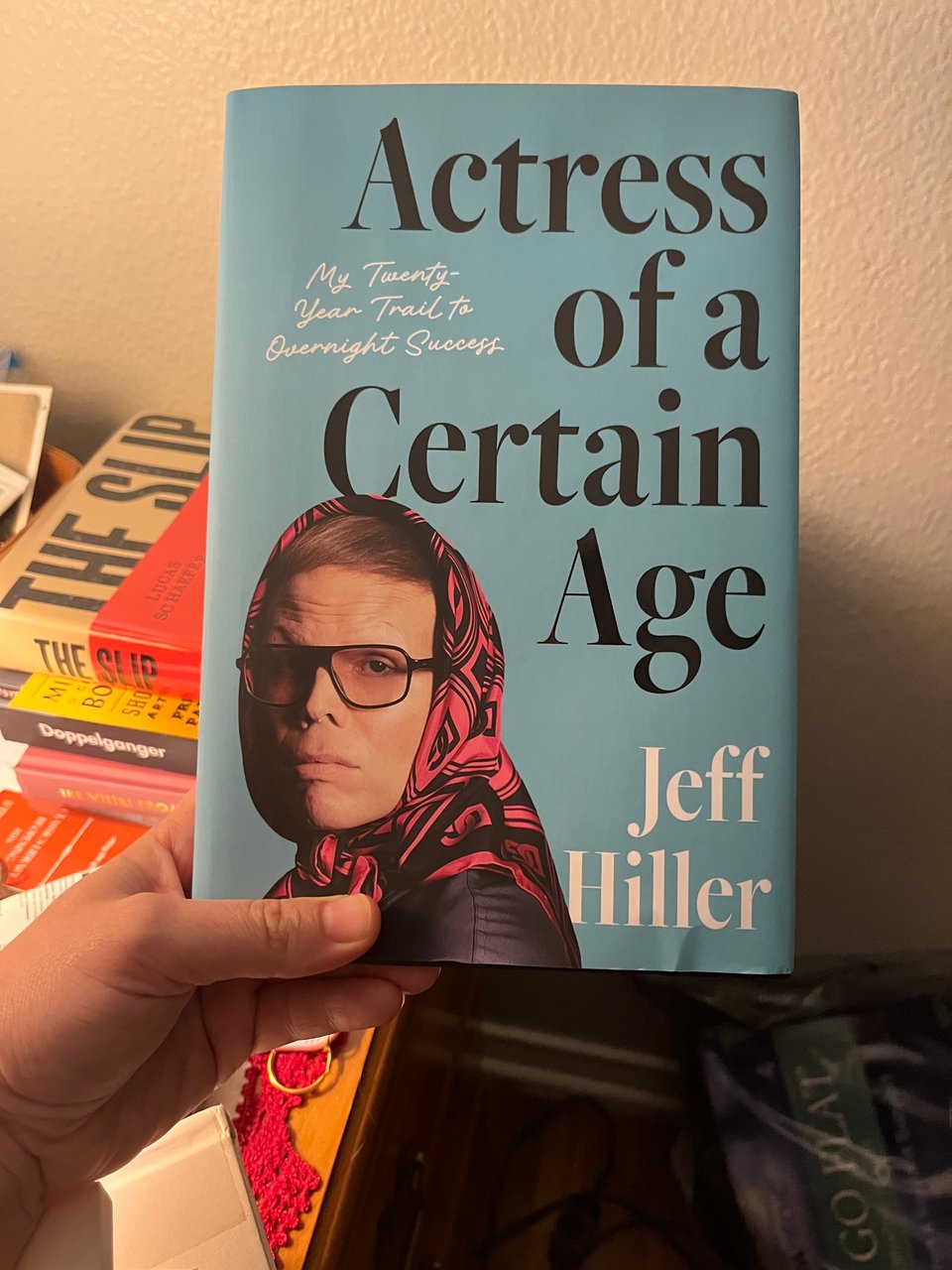 The hardcover of "Actress of a Certain Age" posed in front of a bedside nightstand, which has more books piled up in the background.
