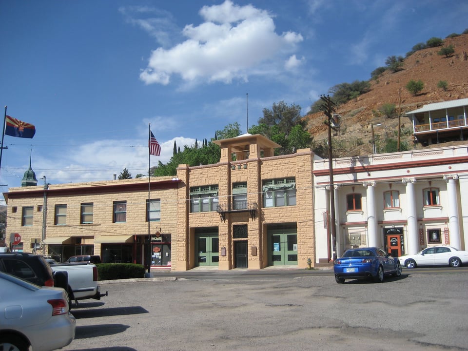 Two buildings in a row - one of amber stone with a U.S. flag and the other whitewashed, on the far side of a parking lot. There is a hill behind them, dotted with shrubs.
