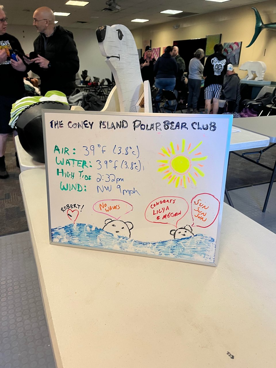 A dry erase board on a folding table in a meeting room. The board says: The Coney Island Polar Bear Club Air: 39 F (3.8 C) Water: 39 F (3.8 C) High Tide: 2:32 p.m. Wind: NW 9 mph. There is an illustartion of two bears swimming on the bottom.