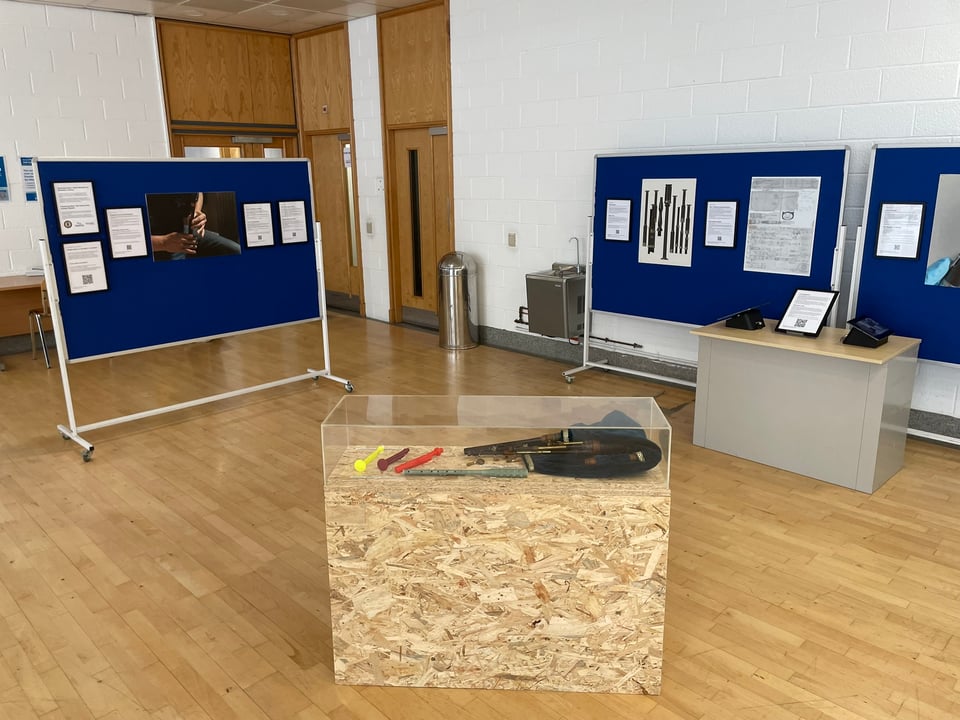Exhibition boards with images and text (not readable as at a distance), and in the foreground an OSB plinth with perspex vitrine cover, containing on the right the early MacAskill set of Lindsay System smallpipes, and on the left four colourful early 3D printed prototypes.