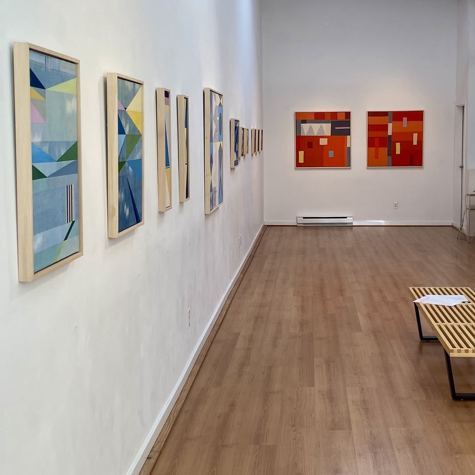 Long view of Tristesse Seeliger's solo exhibition at Deluge Contemporary Art. A row of vertically oriented geometric paintings in blues, greens, and yellows runs along the left wall; two square paintings in reds and oranges hang together on the back wall. Light wood floor and a wooden slat bench in the foreground.