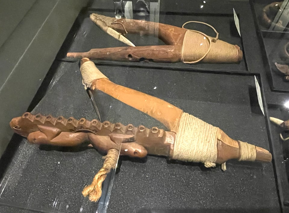 Two wooden Pacific halibut hooks in a museum case. The nearer one is clearly made of two different kinds of wood lashed together to form a 'Y' shape with a metal, inward-facing barb. Carvings of sea otters and an octopus are visible on the nearest wooden arm.