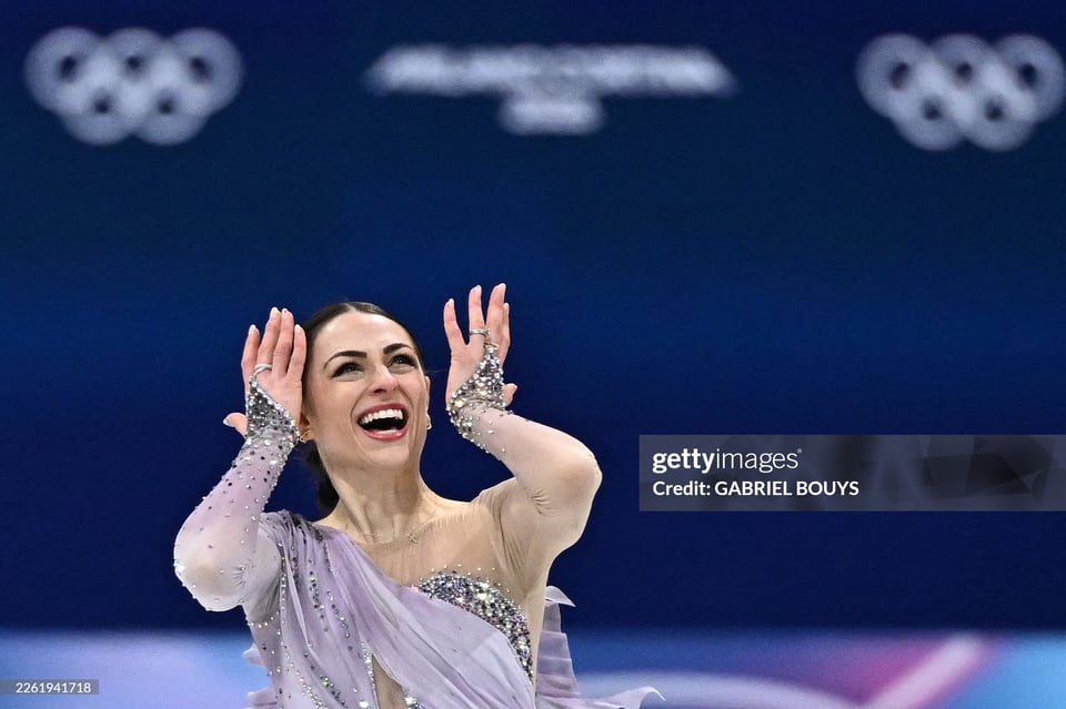 Photo by Figure skater with hands raised in the air, wearing a lavender embellished costume, smiling widely, celebrating during a competition, with Olympic rings visible in the background.