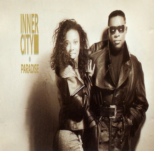 A light-sepia toned photograph of a Black woman and a Black man. They look at the camera. She has long black hair and is turned with her hip towards us and has one arm around his back and the other on his arm. She wears a leather jacket. She has a look of surprise on her face. He has short black hair, sunglasses, and wears a long black leather trenchcoat, with a black shirt underneath. He looks stoic. It is a very MATRIX look, which wouldn't come out for another 10 years.