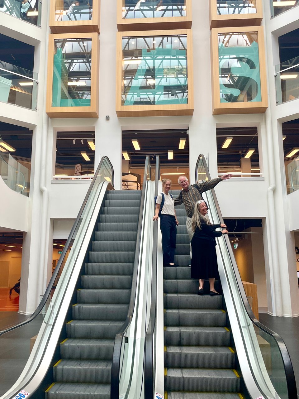 Three people ascend an escalator and look at the camera down below.