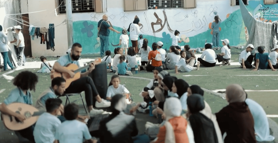 People sitting on the ground, in front of a man playing guitar and someone playing an oud. In the background, more people sitting on the ground and people standing near a painted wall