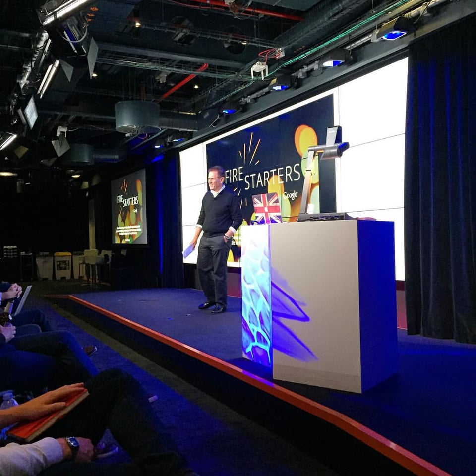 A man stands on stage, presenting to an audience. On the screen behind him is a slide that says FIRESTARTERS.