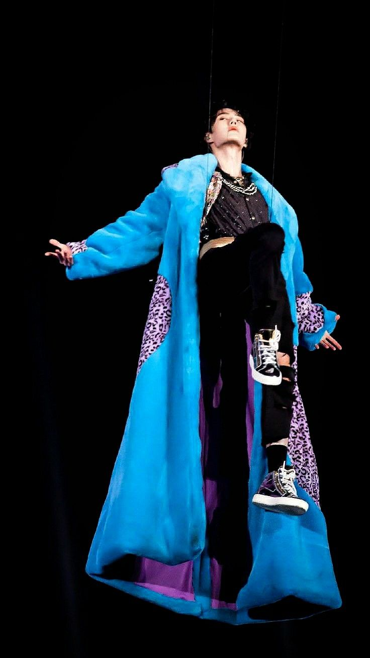 Yibo in mid-air in his famous cookie-monster Nike cape for his new year's eve performance of Rules of My World, 12/31/2020. His arms are outstretched in a jewel-blue coat with a bright purple inner lining and pink leopard-spot patches on the sides. His legs are lifted, bent at the knees, and he's wearing a black sparkly chanel top with gold braided necklaces, a gold belt, ripped black jeans, and black-and-purple tennis shoes. He's suspended by cables and looking up triumphantly off to his right. A king descending.