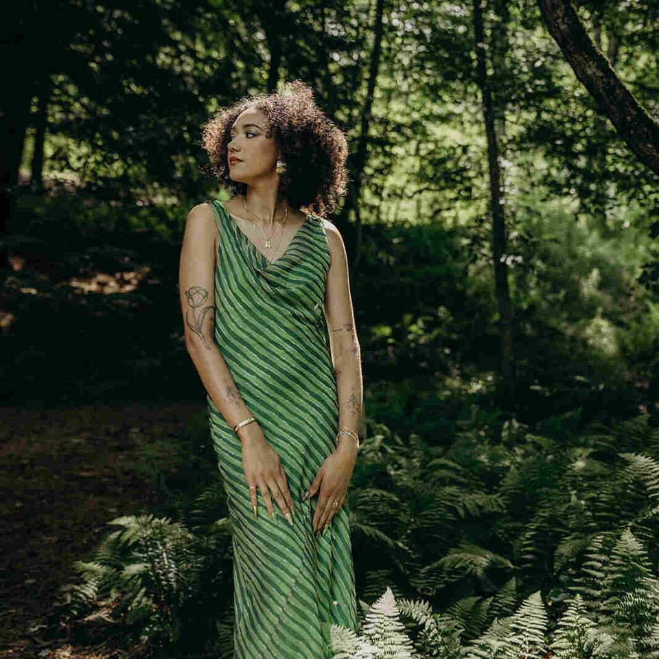 A woman wearing a sleeveless green dress with diagonal stripes stands in the woods with her hands on her legs