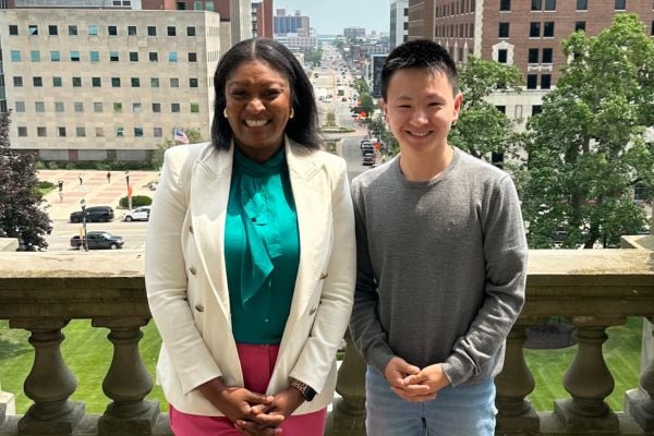 A person in a white blazer and green blouse stands beside a JMC student in a gray sweater, both smiling at the camera. They are positioned on an outdoor balcony overlooking a city street with buildings, trees, and vehicles visible below.