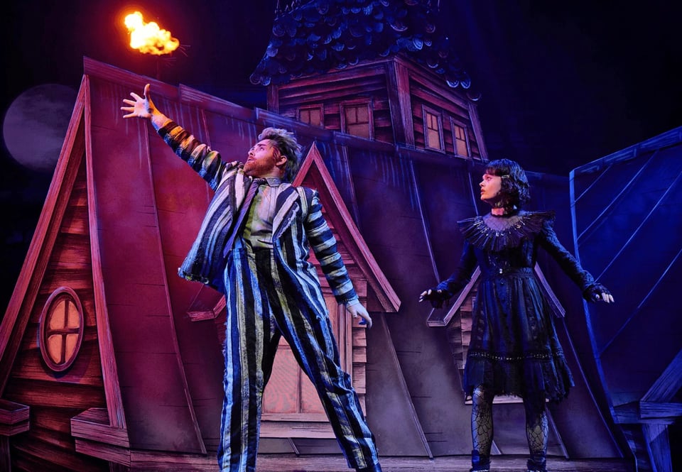 A performer in a striped outfit dramatically gestures with a flame, while another performer in a dark dress watches on a mystical, theatrical set.