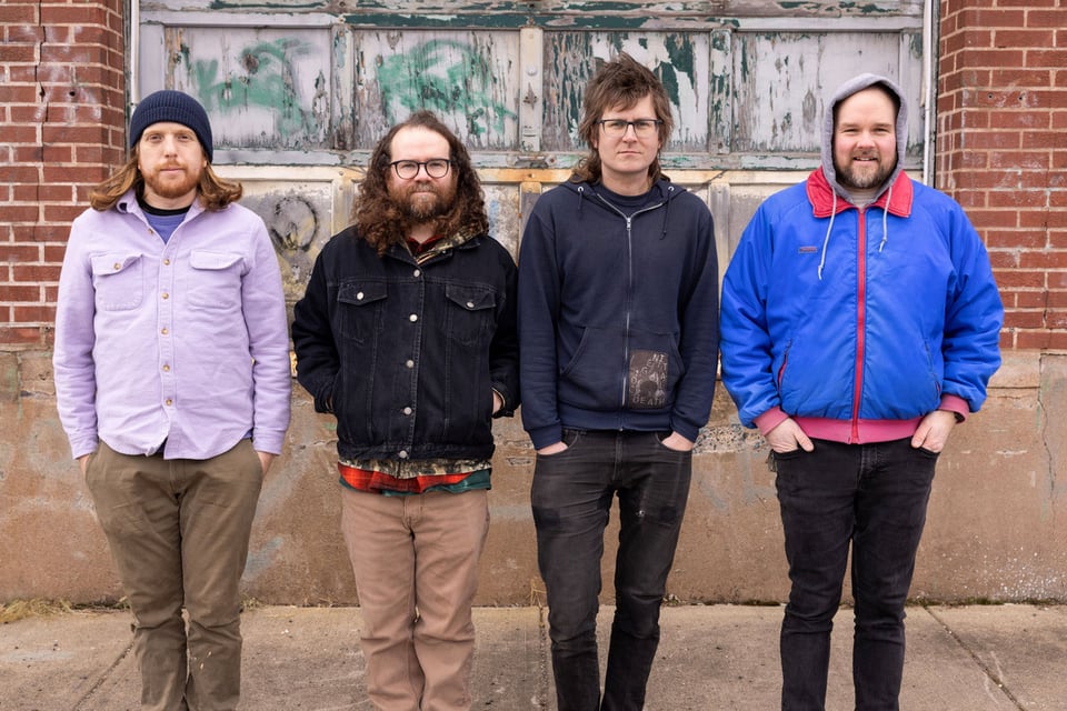 Four men stand in front of a boarded up window set in a brick wall.