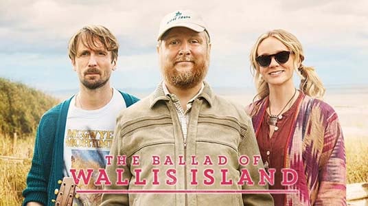 Photo: Three people standing in a grassy field under an overcast sky. Left: man with brown hair and beard wearing a white t-shirt with blue sleeves. Centre: bearded man in a beige baseball cap and tan multi-pocket vest over a collared shirt, smiling broadly. Right: blonde woman with plaited hair wearing sunglasses and a striped burgundy and cream shirt. Text overlay at bottom reads "THE BALLAD OF WALLIS ISLAND" in pink serif lettering.