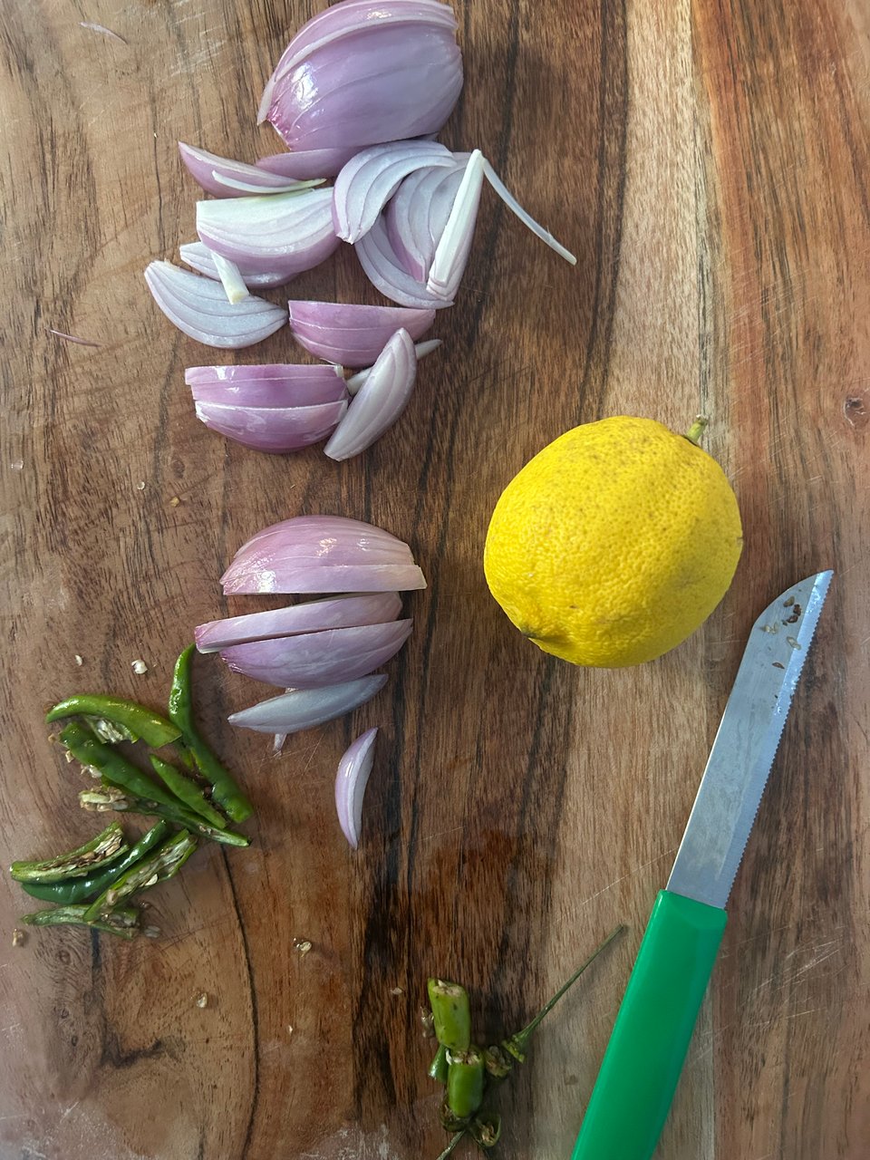 a close up of a cutting board with chopped shallots, thai chili peppers, a whole lemon, and a small green cutting knife