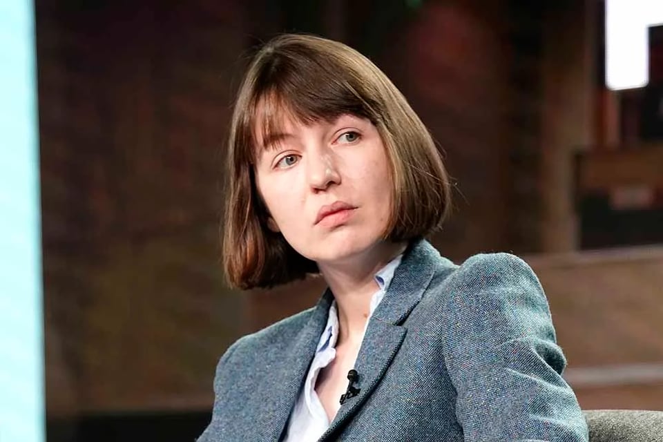 Sally ROoney, wearing a gray blazer, brown bob haircut and bangs, looking to her left at an event