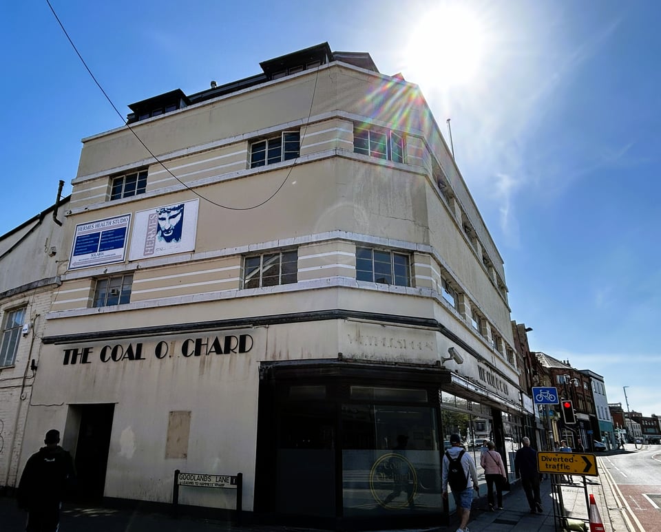 A three storey art Deco building with a later mansard addition behind the parapet. The building is cream, decorative lines in white and Crittall windows set between white concrete bands. Some lettering, using a Cotton Club style font, are still on it from when it was a pub.