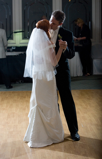 Photo of a white cis couple kissing at their wedding. A red-haired woman is in a white gown with headpiece, and the taller brown-haired man is in a tuxedo w/white tie