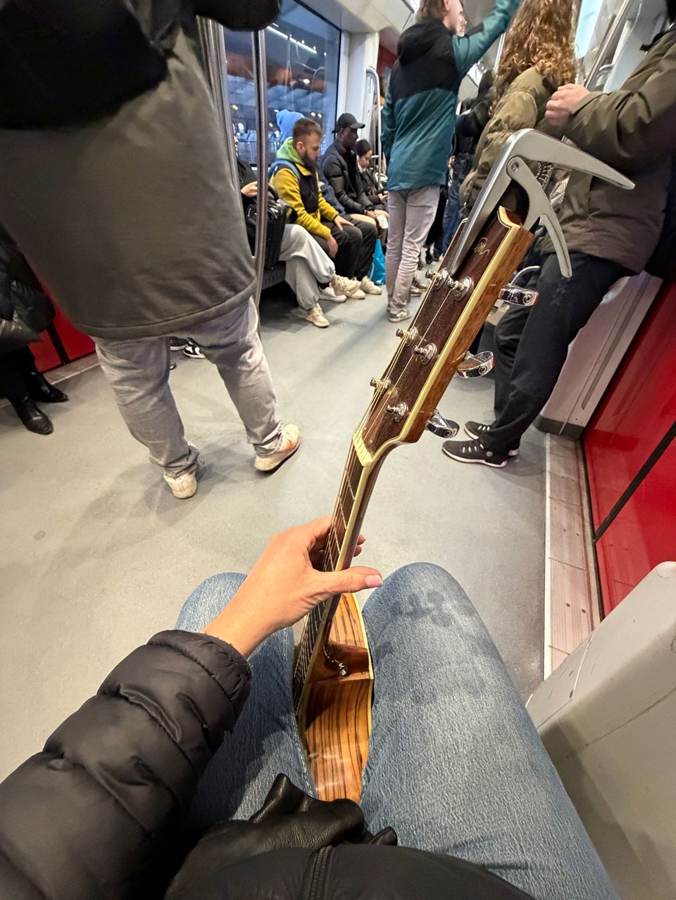 A guitar is held between two legs of a woman sitting on a metro.